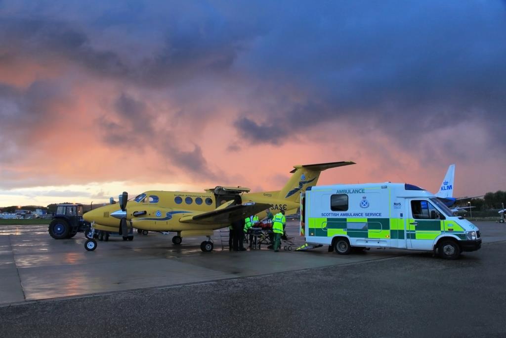 NHS Scotland staff and paramedics undertake an emergency patient transfer between two emergency service vehicles - a small yellow painted Scottish Ambulance Service twin-propelled aircraft and an ambulance - against a backdrop of dark stormy skies over a tarmac airfield. image